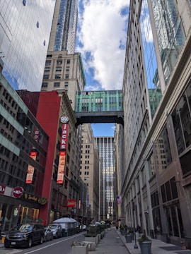 A city street with tall buildings on both sides. A skybridge connects two buildings at an upper level near the top of the frame. The buildings are a mix of modern glass and older styles; one building has a red façade with signs advertising a 99 cent store and a restaurant named Jacks. Several vehicles including cars and vans are parked along the street. There are a few pedestrians on the sidewalks.