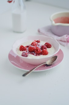 A bowl of creamy porridge topped with fresh raspberries is set on a pink plate. A spoon rests beside the bowl, and there is a light sprinkling of what appears to be shredded coconut. In the background, a bottle of milk and a light pink pot are visible, contributing to a soft and harmonious setting.
