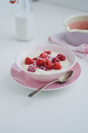 A bowl of creamy porridge topped with fresh raspberries is set on a pink plate. A spoon rests beside the bowl, and there is a light sprinkling of what appears to be shredded coconut. In the background, a bottle of milk and a light pink pot are visible, contributing to a soft and harmonious setting.