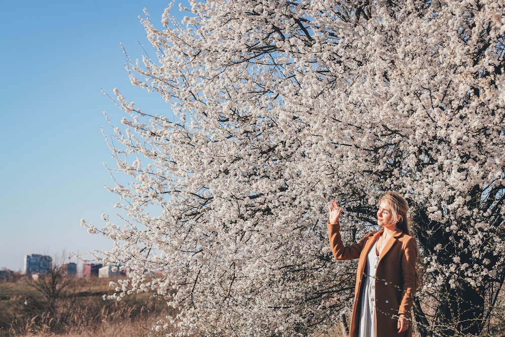 woman in brown coat standing near white cherry blossom tree during daytime