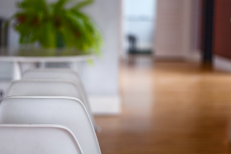 A row of white chairs is aligned next to a table, with a green plant placed on the table. The background reveals a wooden floor extending into a hallway, with walls in soft colors and slight blurriness, creating a feeling of depth.