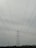 A large metal electricity transmission tower stands amidst a landscape, towering over a series of power lines stretching wide across the frame. The horizon features a slight silhouette of hills in the distance under a cloudy sky.