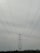 A large metal electricity transmission tower stands amidst a landscape, towering over a series of power lines stretching wide across the frame. The horizon features a slight silhouette of hills in the distance under a cloudy sky.