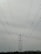 A large metal electricity transmission tower stands amidst a landscape, towering over a series of power lines stretching wide across the frame. The horizon features a slight silhouette of hills in the distance under a cloudy sky.