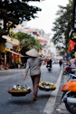 A person in a conical hat carrying produce in baskets on a city street