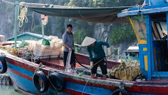 Two men are on a wooden fishing boat, with one of them using a hose to wash the deck. The boat is filled with various fishing gear such as ropes, nets, and crates. The backdrop consists of rocky cliffs and greenery, suggesting a coastal environment.