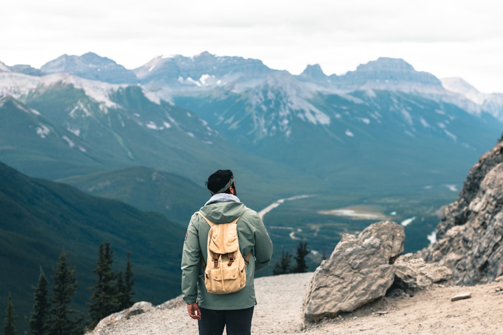 man in brown jacket and black pants standing on rock formation looking at mountains during daytime