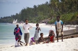 A group of children and a woman are on a white sandy beach with tropical greenery and palm trees in the background. The children are dressed in casual summer clothes and are either standing, sitting, or playing with sand. In the background, there are people walking and sailing boats in the turquoise water.