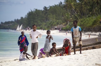 A group of children and a woman are on a white sandy beach with tropical greenery and palm trees in the background. The children are dressed in casual summer clothes and are either standing, sitting, or playing with sand. In the background, there are people walking and sailing boats in the turquoise water.