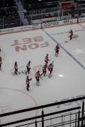 A group of ice hockey players wearing red and white uniforms are gathered on an indoor rink. They appear to be communicating or celebrating, holding their sticks upright. The rink is marked with various advertisements and logos, and there is a seating area for spectators.