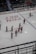 A group of ice hockey players wearing red and white uniforms are gathered on an indoor rink. They appear to be communicating or celebrating, holding their sticks upright. The rink is marked with various advertisements and logos, and there is a seating area for spectators.