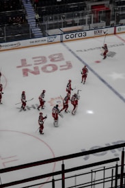 A group of ice hockey players wearing red and white uniforms are gathered on an indoor rink. They appear to be communicating or celebrating, holding their sticks upright. The rink is marked with various advertisements and logos, and there is a seating area for spectators.