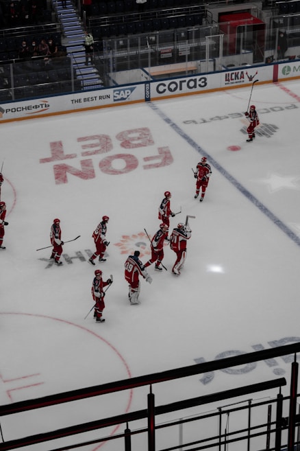 A group of ice hockey players wearing red and white uniforms are gathered on an indoor rink. They appear to be communicating or celebrating, holding their sticks upright. The rink is marked with various advertisements and logos, and there is a seating area for spectators.