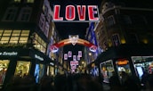 A vibrant street scene at night features a large illuminated sign spelling out 'LOVE' suspended above a bustling shopping area. Surrounding buildings are adorned with festive lights and decorations, and people are visible walking along the street. A secondary sign reads 'Carnaby Christmas Revolution @CarnabyLondon'.