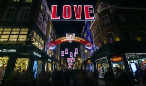 A vibrant street scene at night features a large illuminated sign spelling out 'LOVE' suspended above a bustling shopping area. Surrounding buildings are adorned with festive lights and decorations, and people are visible walking along the street. A secondary sign reads 'Carnaby Christmas Revolution @CarnabyLondon'.