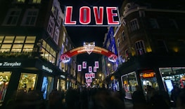 A vibrant street scene at night features a large illuminated sign spelling out 'LOVE' suspended above a bustling shopping area. Surrounding buildings are adorned with festive lights and decorations, and people are visible walking along the street. A secondary sign reads 'Carnaby Christmas Revolution @CarnabyLondon'.