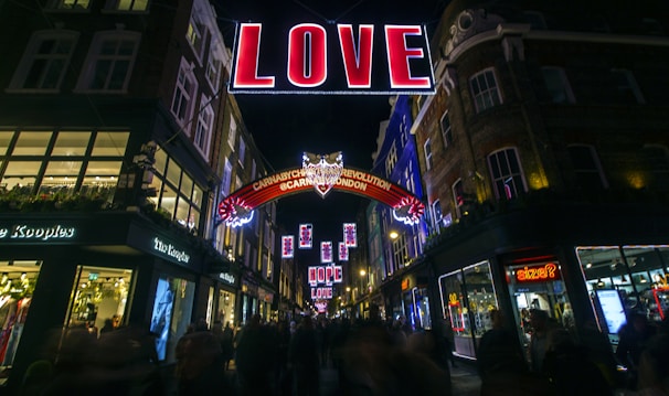 A vibrant street scene at night features a large illuminated sign spelling out 'LOVE' suspended above a bustling shopping area. Surrounding buildings are adorned with festive lights and decorations, and people are visible walking along the street. A secondary sign reads 'Carnaby Christmas Revolution @CarnabyLondon'.