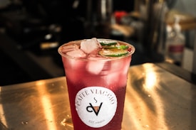 A clear plastic cup filled with a red beverage sits on a metal surface. The drink is topped with ice cubes and slices of lime. The cup has a label that reads 'Caf&eacute; Viaggio.' The background is blurred, suggesting an indoor setting with lighting that creates a warm ambiance.