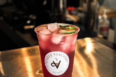 A clear plastic cup filled with a red beverage sits on a metal surface. The drink is topped with ice cubes and slices of lime. The cup has a label that reads 'Café Viaggio.' The background is blurred, suggesting an indoor setting with lighting that creates a warm ambiance.