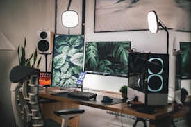 A modern workspace setup featuring dual monitors with plant-themed screens, a mechanical keyboard, a smartphone stand, and a gaming chair. The ambient lighting includes two circular studio lights on stands. Additional elements include a speaker, a computer tower with illuminated fans, and decorative small plants on the desk.