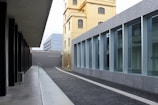 A modern architectural scene with sleek lines and geometric shapes. A tall, yellowish building stands adjacent to a structure with large glass windows framed in gray. The foreground features a pathway paved with dark bricks, leading towards more contemporary buildings in the background. There is an interplay of old and new architectural styles.