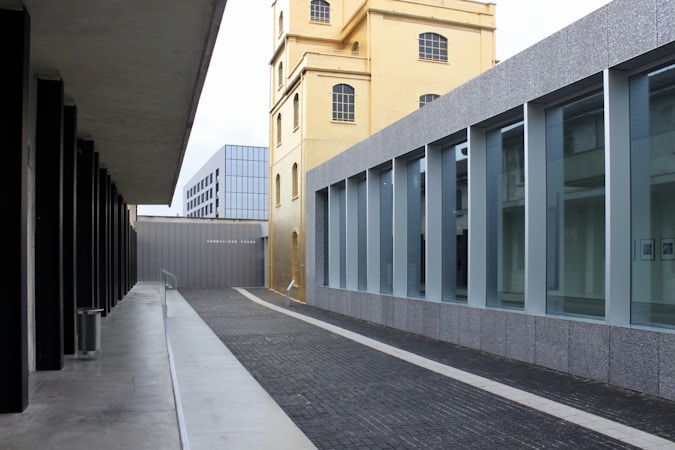A modern architectural scene with sleek lines and geometric shapes. A tall, yellowish building stands adjacent to a structure with large glass windows framed in gray. The foreground features a pathway paved with dark bricks, leading towards more contemporary buildings in the background. There is an interplay of old and new architectural styles.
