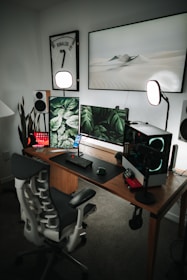 A modern and stylish home office setup with a wooden desk featuring multiple monitors displaying green leaf patterns and a sleek gaming chair. The setup includes speakers, a microphone, and a CPU with glowing fans. The walls are adorned with a framed sports jersey and minimalist art, while soft lighting and a potted plant add ambiance.