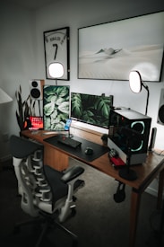 A modern and stylish home office setup with a wooden desk featuring multiple monitors displaying green leaf patterns and a sleek gaming chair. The setup includes speakers, a microphone, and a CPU with glowing fans. The walls are adorned with a framed sports jersey and minimalist art, while soft lighting and a potted plant add ambiance.