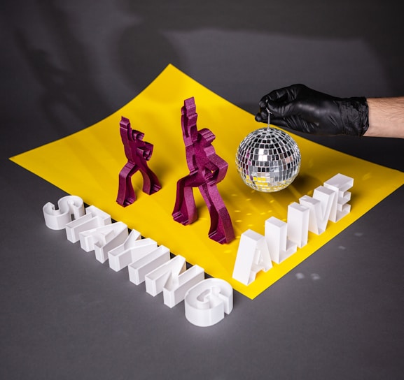 Two geometric, purple figures resembling dancers are placed on a bright yellow surface. Large white 3D letters forming the words 'STAYING ALIVE' are positioned in front of them. A hand wearing a black glove holds a small disco ball above the scene. The yellow surface contrasts with the surrounding dark background.