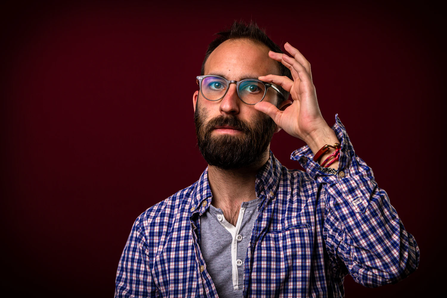 A bearded man in a checked shirt adjusting heavy dark frames against a deep red wall