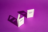 Two white book-like objects stand upright on a vibrant purple background. The object on the left has the word 'SHIT' and casts a shadow that clearly mirrors the text. The object on the right reads 'DIFFERENT SHIT' and similarly projects a shadow with those words.