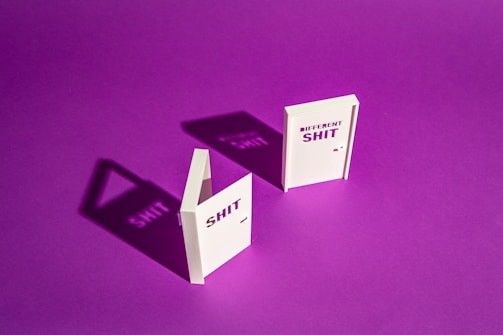 Two white book-like objects stand upright on a vibrant purple background. The object on the left has the word 'SHIT' and casts a shadow that clearly mirrors the text. The object on the right reads 'DIFFERENT SHIT' and similarly projects a shadow with those words.