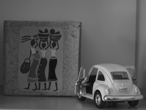 A black and white photograph features a small scale model of a vintage car with its doors open. Next to the car, there is a textured surface with a decorative design depicting three abstract figures, each carrying pots on their heads.