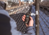 A person holding a digital audio recorder close to a chain-link fence with a blurred background depicting a road and buildings.