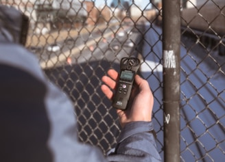 A person holding a digital audio recorder close to a chain-link fence with a blurred background depicting a road and buildings.