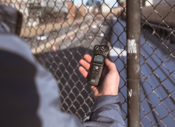 A person holding a digital audio recorder close to a chain-link fence with a blurred background depicting a road and buildings.