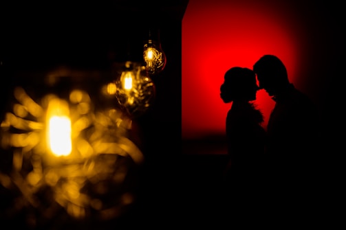 Silhouetted profile of a couple facing each other against a striking red background. On the left side, there are several warm, glowing light bulbs with intricate filament designs creating a soft focus effect.