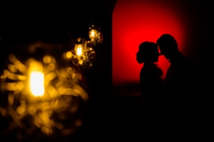Silhouetted profile of a couple facing each other against a striking red background. On the left side, there are several warm, glowing light bulbs with intricate filament designs creating a soft focus effect.