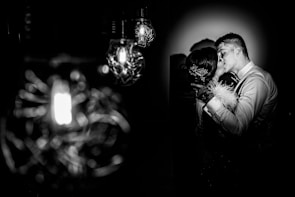 A romantic black and white photograph featuring a couple sharing an intimate kiss. The man, dressed in a formal shirt, embraces the woman, who has a floral hair accessory and a feather detail on her clothing. The dimly lit setting is enhanced by several ornate lightbulbs, creating a dramatic and intimate atmosphere.