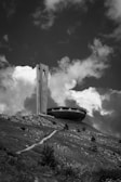 A large concrete structure resembling a flying saucer sits on a grassy hill under a partly cloudy sky. A tall tower with a star emblem is adjacent to the saucer-like building. The foreground features a path leading up to the structure, surrounded by rough terrain and sparse vegetation.