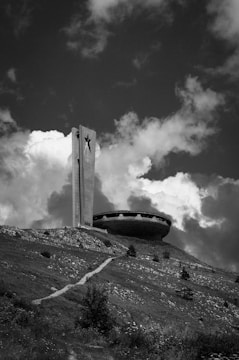 A large concrete structure resembling a flying saucer sits on a grassy hill under a partly cloudy sky. A tall tower with a star emblem is adjacent to the saucer-like building. The foreground features a path leading up to the structure, surrounded by rough terrain and sparse vegetation.