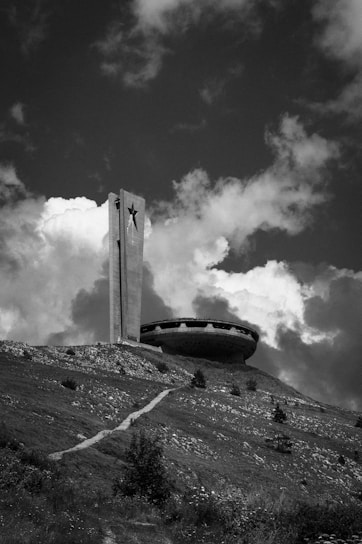 A large concrete structure resembling a flying saucer sits on a grassy hill under a partly cloudy sky. A tall tower with a star emblem is adjacent to the saucer-like building. The foreground features a path leading up to the structure, surrounded by rough terrain and sparse vegetation.