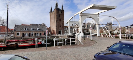 A picturesque scene featuring a historic brick tower with pointed spires beside a drawbridge spanning a narrow canal. Traditional Dutch architecture includes buildings with gabled roofs and a moored boat with a red cover along the waterway. The cobblestone foreground leads the eyes to the idyllic setting under a partly cloudy sky.