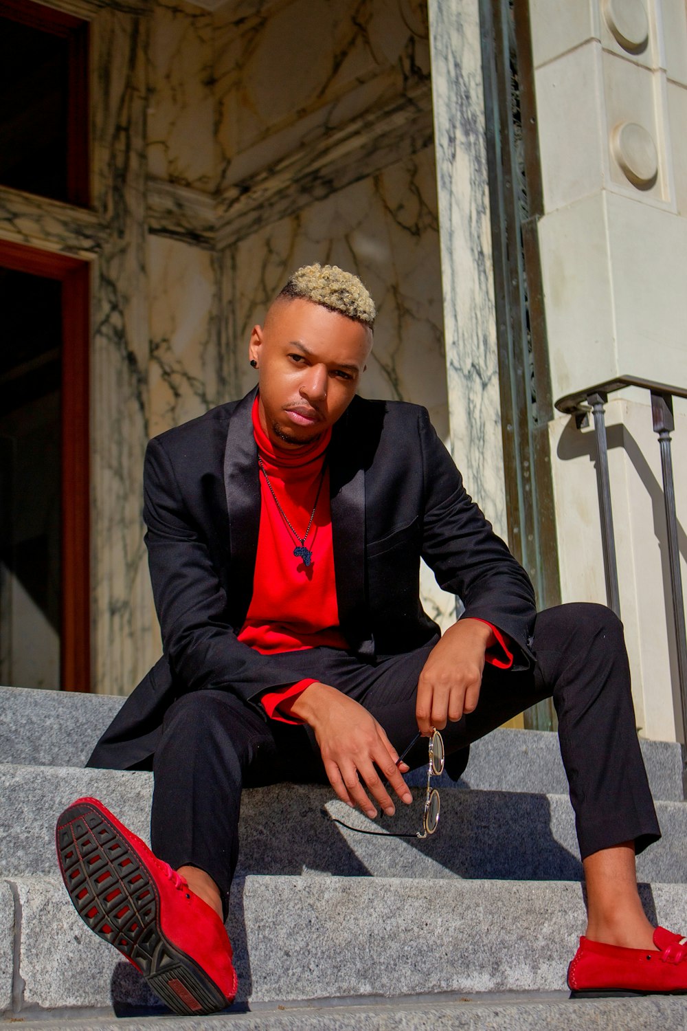 man in black suit jacket and red dress shirt sitting on gray concrete stairs