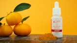 A small white bottle labeled 'LUFF CBD Tincture' sits on a surface next to a pile of turmeric powder. Three bright orange fruits with green leaves are also present, with a vibrant orange background enhancing the composition.