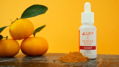 A small white bottle labeled 'LUFF CBD Tincture' sits on a surface next to a pile of turmeric powder. Three bright orange fruits with green leaves are also present, with a vibrant orange background enhancing the composition.