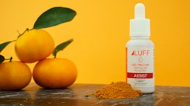 A small white bottle labeled 'LUFF CBD Tincture' sits on a surface next to a pile of turmeric powder. Three bright orange fruits with green leaves are also present, with a vibrant orange background enhancing the composition.