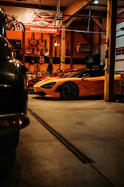 A dimly lit garage features a bright orange sports car prominently in the foreground. The garage walls are adorned with several guitars, and a large Budweiser sign hangs above. To the left, part of another vehicle is visible, partially shrouded in shadow.