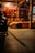 A dimly lit garage features a bright orange sports car prominently in the foreground. The garage walls are adorned with several guitars, and a large Budweiser sign hangs above. To the left, part of another vehicle is visible, partially shrouded in shadow.
