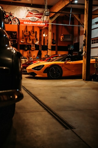 A dimly lit garage features a bright orange sports car prominently in the foreground. The garage walls are adorned with several guitars, and a large Budweiser sign hangs above. To the left, part of another vehicle is visible, partially shrouded in shadow.
